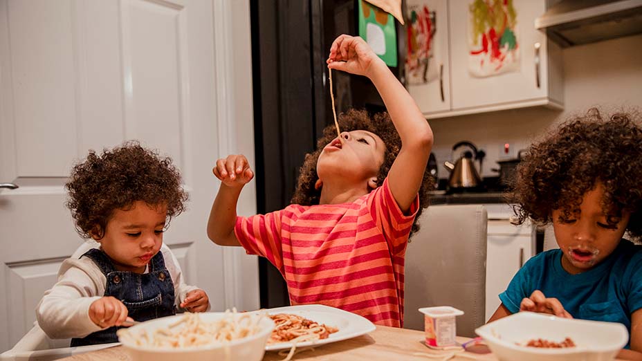 Three young children round a table eating spaghetti.One child dangles spaghetti into his mouth