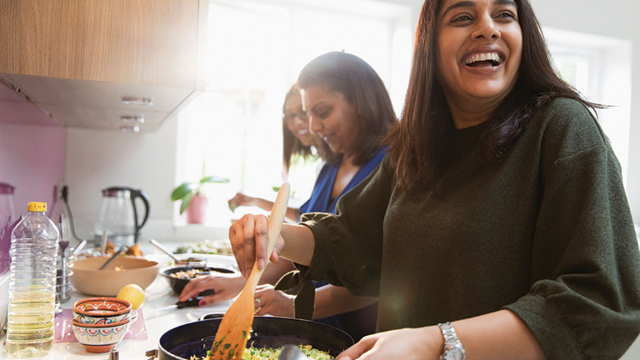 Happy women laughing whilst cooking food at stove in kitchen